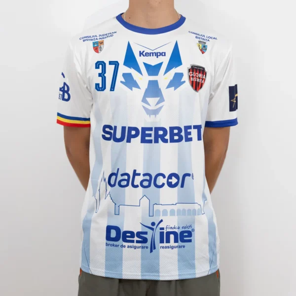 Tricou de joc handbal Gloria Bistrița – Alb (față) Tricou de joc handbal Gloria Bistrița, alb cu accente albastre, văzut din față — sigla Kempa, stema clubului și sponsorii
